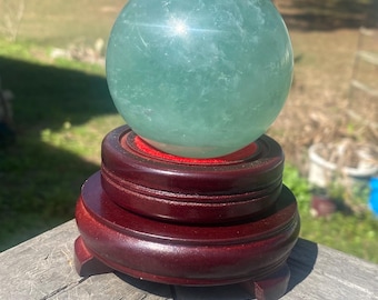 May include: A light green crystal sphere rests on a tiered, dark reddish-brown wooden stand. The sphere is translucent and reflects light, with a red felt lining on the stand. The piece has a natural and elegant appearance.