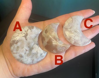 May include: Three polished agate moon-shaped stones in varying sizes and colours. The largest is a full moon shape, while the others are crescent moons. The stones display a mix of white, grey, and brown hues, with unique patterns.