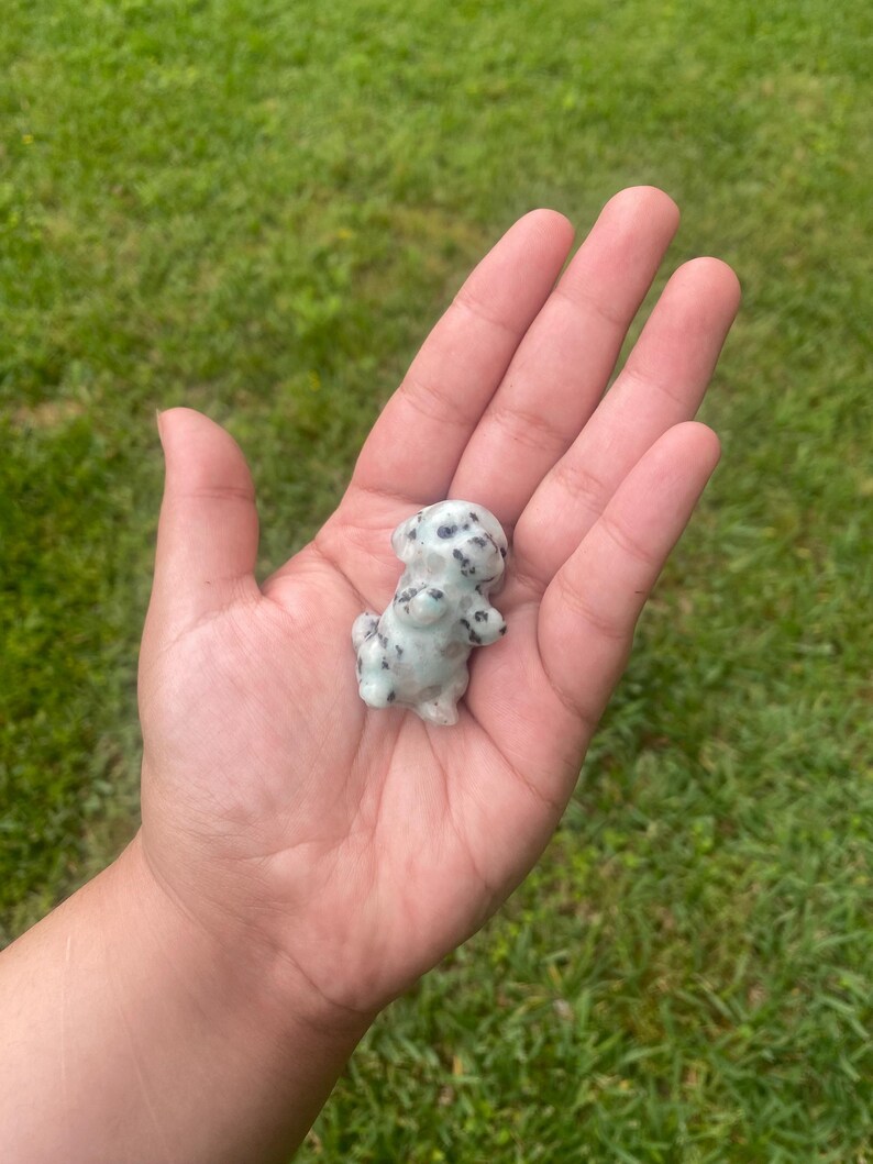 May include: A small, light-coloured figurine of a Dalmatian dog with black spots. The dog is standing upright with its front paws together. The figurine is held in an open hand against a backdrop of green grass.
