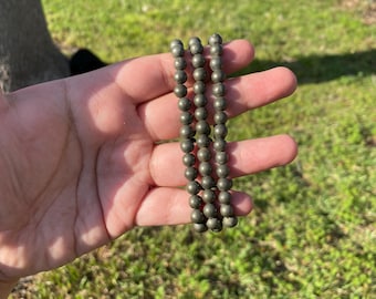 May include: Three strands of small, round, metallic-looking beads. The beads are a dark, muted colour, possibly grey or bronze. The strands are held in a hand against a background of green grass and a tree trunk.