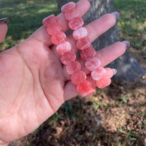 May include: Two strands of pink and red gemstone bracelets. Each bracelet is composed of rounded, polished stones. The stones vary in color from light pink to deep red. The bracelets are held in a hand.