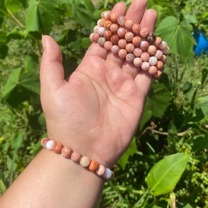 May include: A collection of beaded bracelets in shades of peach, brown, and white. The bracelets are made of round, polished stones and are displayed on a hand and wrist against a backdrop of green foliage.