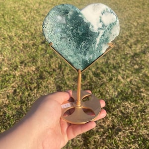 May include: A heart-shaped stone with green and white patterns, displayed on a gold-colored metal stand. The stone is held in place by a thin, angled support. The stand has a circular base with a cross-shaped design.