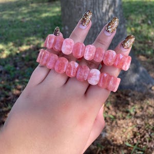 May include: Two pink gemstone bracelets, each composed of rectangular beads. The bracelets are displayed on a hand, with a blurred background of green grass and a tree trunk. The gemstones have a translucent quality.