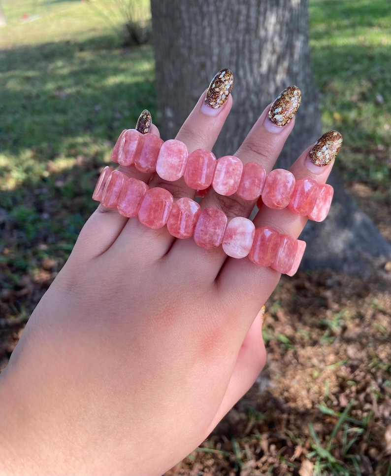 May include: Two pink gemstone bracelets, each composed of rectangular beads. The bracelets are displayed on a hand, with a blurred background of green grass and a tree trunk. The gemstones have a translucent quality.