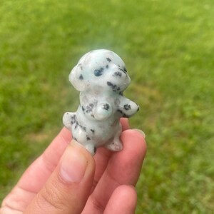 May include: A small, light green dog figurine with black spots. The dog is standing upright with its arms outstretched. The figurine is made of a speckled stone material.