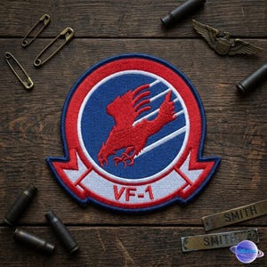 May include: Embroidered VF-1 patch featuring a red phoenix design on a blue background, encircled by a red and white border. The patch is surrounded by brass safety pins, bullets, and name tags on a wooden surface.