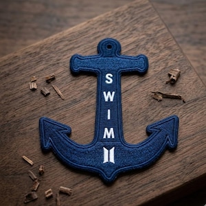 May include: A navy blue embroidered anchor patch with the word "SWIM" in white letters. The patch is on a wooden surface with small wood shavings scattered around it. The anchor design features a classic shape.