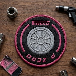 May include: Embroidered patch featuring the Pirelli logo and a stylized wheel design. The patch is black with pink and gray accents, and the text "P ZERO" is visible. The patch is round and likely for apparel or accessories.