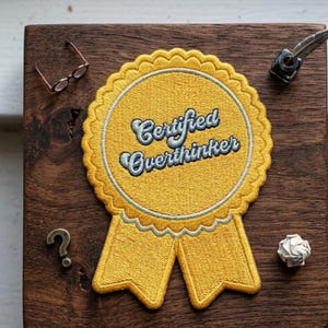 May include: A yellow embroidered patch with the words "Certified Overthinker" in a whimsical font. The patch is on a dark wooden surface, with small reading glasses, a question mark, and an inkwell nearby.