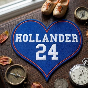 May include: A blue heart-shaped patch with the word "HOLLANDER" and the number "24" in white. The patch has an orange border. Wooden clogs, compasses, a stopwatch, and a coin are arranged around the patch on a wooden surface.