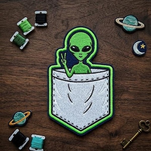 May include: Embroidered patch featuring a green alien giving a peace sign, emerging from a white pocket with a green border. Additional patches include a planet, moon, and spools of thread. A vintage key is also present.