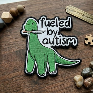 May include: Embroidered patch featuring a green dinosaur holding a knife in its mouth. The patch has the words "fueled by autism" in white lettering with a black border. The patch is on a wooden surface.