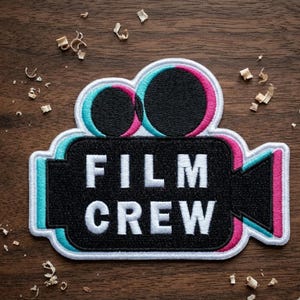 May include: A black and white embroidered patch in the shape of a film camera with the words "FILM CREW" in white. The camera lens has a teal and pink outline. The patch is on a wooden surface with wood shavings.