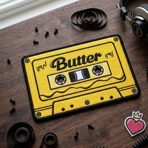 May include: Yellow embroidered patch in the shape of a cassette tape with the word "Butter" in black script. The patch has black trim and is on a wooden surface. Other items include screws, headphones, and a heart patch.