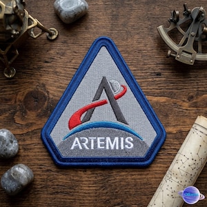 May include: A triangular patch with a blue border and the word "ARTEMIS" in white. The patch features a gray "A" with a red and white swoosh, and a blue arc. The patch is on a wooden surface.
