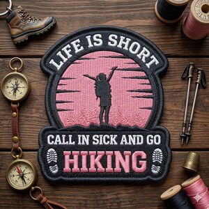 May include: Embroidered patch with the text "LIFE IS SHORT CALL IN SICK AND GO HIKING" featuring a hiker silhouette against a pink sunset. The patch is surrounded by hiking gear, including a compass, hiking boots, and trekking poles.