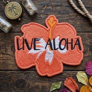 May include: Embroidered patch in the shape of a hibiscus flower, with the words "LIVE ALOHA" in black lettering. The flower is orange with white and yellow accents. The patch is on a wooden surface with a compass and rope.