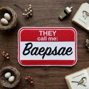 May include: A rectangular patch with a red border and a white center. The text "THEY call me: Baepsae" is embroidered in black. The patch is on a wooden surface with bird nests, eggs, and a whistle.