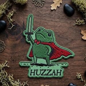 May include: Embroidered patch featuring a green frog in a red cape, holding a sword. The patch has the word "Huzzah" at the bottom. The frog has a red bow tie. The patch is surrounded by decorative elements on a wooden surface.