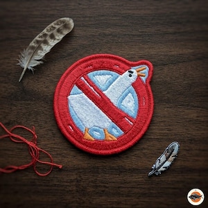 May include: Embroidered patch featuring a seagull inside a red circle with a red slash, indicating a prohibition. The patch is surrounded by a feather and red thread on a wooden surface. The seagull is white and blue.