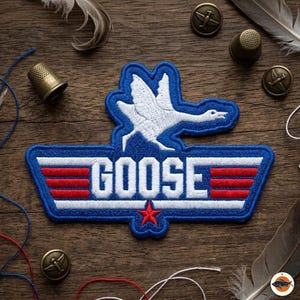 May include: Embroidered patch featuring a white goose in flight, above the word "GOOSE" in white block letters. The patch has a blue border with red and white stripes and a red star. The patch is on a wooden surface.