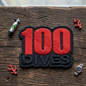 May include: A black and red embroidered patch with the text "100 DIVES". The patch is surrounded by a miniature scuba tank, a wristwatch, and coral decorations, all set on a wooden surface.