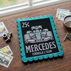 May include: A black patch with a teal border, featuring a Formula 1 race car design. The patch reads "MERCEDES FORMULA 1 TEAM" and "25¢". Additional details include a small checkered flag and postage stamps.