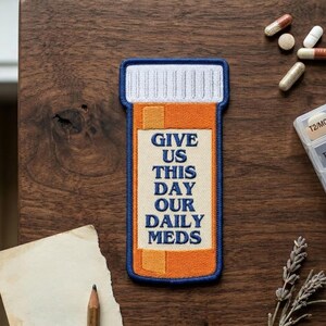 May include: Embroidered patch in the shape of a prescription pill bottle, with the words "GIVE US THIS DAY OUR DAILY MEDS" in navy blue. The bottle is orange with a white and blue cap. Several pills and a pill organizer are in the background.