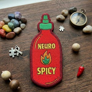 May include: A red embroidered patch in the shape of a hot sauce bottle with the words "NEURO SPICY" and a flame design. The patch is surrounded by small stones, a compass, and a miniature red chili pepper.