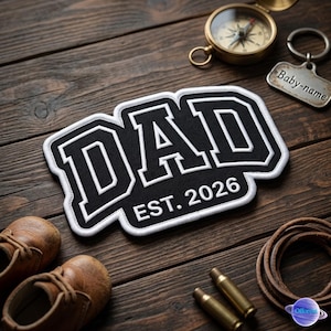May include: A black and white embroidered patch with the word "DAD" and "EST. 2026". The patch is surrounded by a white border. Also shown are a compass, a keychain that says "Baby-name", and small brown leather shoes.