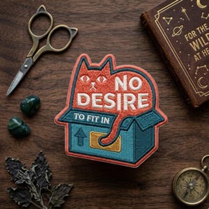 May include: Embroidered patch featuring a red cat inside a blue box with the words "NO DESIRE TO FIT IN". The patch is surrounded by a pair of cat-shaped scissors, green stones, a small compass, and a book.