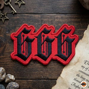 May include: A red and black embroidered patch featuring the number "666" in a gothic font. The patch has a red border and is set against a dark wooden background. The patch is a decorative item.