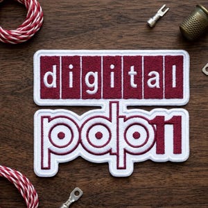 May include: Two embroidered patches with the words "digital" and "pop11" in white lettering on a maroon background. The patches have a white border. The image is on a wooden surface with sewing notions and red and white twine.