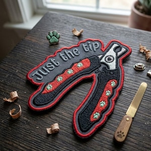 May include: An embroidered patch with the text "Just the tip" above a pet nail clipper design. The patch is black, red, and gray with green paw prints. A small green paw print and a nail file with a wooden handle are also visible.
