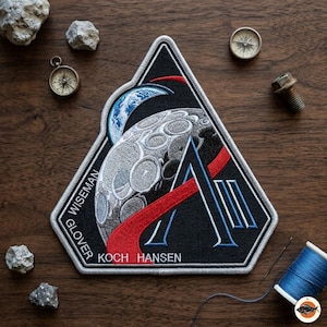 May include: Embroidered patch featuring a detailed depiction of the moon, Earth, and a stylized letter "A" against a black background. The patch includes the names "WISEMAN", "GLOVER", "KOCH", and "HANSEN".
