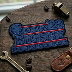 May include: Embroidered patch with the words "Clayton Bigsby 28" in blue thread, with red horizontal lines. The phrase "LET THAT HATE OUT!" is stitched below. The patch is on a wooden surface, with a whistle, wrench, and leather keychain.