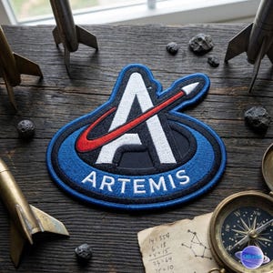 May include: Embroidered Artemis patch with a blue border, white "A" and red swoosh. The word "ARTEMIS" is in white. The patch is on a wooden surface with toy rockets, rocks, and a compass.