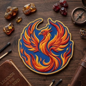 May include: Embroidered patch featuring a phoenix rising from flames. The patch has a blue background with a gold border. The phoenix is depicted in shades of red, orange, and yellow. The patch is surrounded by amber stones, a compass, and a ring.