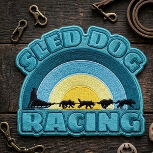 May include: Embroidered patch with the words "SLED DOG RACING" in teal. The design features a sled and dogs racing across a yellow and blue sky. The patch is surrounded by a teal border. Accessories are scattered around the patch.