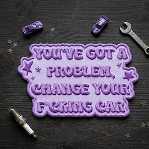 May include: A lavender embroidered patch with the text "YOU'VE GOT A PROBLEM, CHANGE YOUR F*CKING CAR". The patch is surrounded by star shapes and has a decorative border. Two toy cars, a wrench, and a spark plug are also visible.