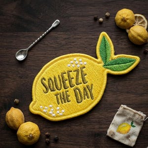 May include: A yellow lemon-shaped embroidered patch with the text "SQUEEZE THE DAY" sits on a dark wood surface. The patch is next to a silver bar spoon, lemons, peppercorns, and a small bag with a lemon illustration.