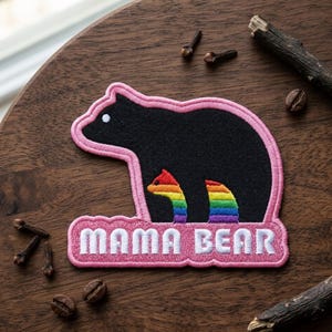 May include: Embroidered patch featuring a black bear silhouette with a pink outline. The bear has a rainbow design and the text "MAMA BEAR" in white on a pink banner. The patch is on a wooden surface.