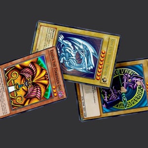 May include: Three collectible Yu-Gi-Oh! trading cards. The cards feature illustrations of a dragon, a magician, and a monster. The cards have gold borders and text. The cards are on a dark gray background.