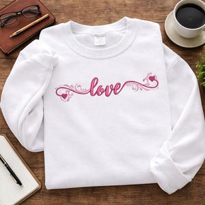 May include: White sweatshirt featuring the word "love" in pink script, embellished with decorative swirls and hearts. The design is centrally placed. The sweatshirt is made of a soft material.