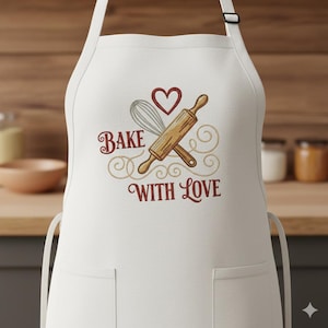 Op de afbeelding: Witte schort met twee voorzakken en een verstelbare nekband. De schort heeft een geborduurd ontwerp met een deegroller, garde en hart, samen met de tekst "Bake With Love". Keuken- en bakthema.