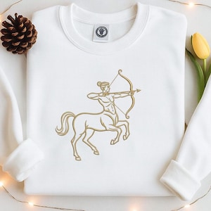 May include: White sweatshirt featuring a gold embroidered Sagittarius zodiac symbol. The design depicts a centaur archer with a bow and arrow. The sweatshirt has a crew neck and long sleeves, perfect for casual wear.
