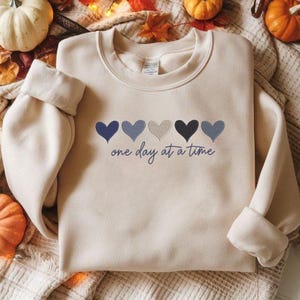May include: A cream-colored sweatshirt with five heart designs in shades of blue and gray. The phrase "one day at a time" is written in a script font below the hearts. The sweatshirt has long sleeves and a crew neck.