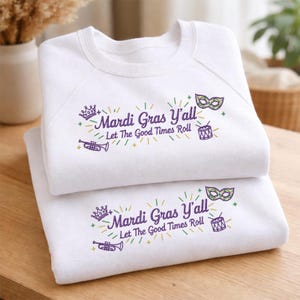 May include: Two white sweatshirts are stacked on a wooden surface. Each sweatshirt has the text "Mardi Gras Y'all Let The Good Times Roll" in purple and green, along with crown, mask, trumpet, and drum graphics.