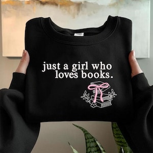May include: Black sweatshirt with the text "just a girl who loves books." A graphic of stacked books with a pink bow and floral accents is below the text. The sweatshirt is held by a person.